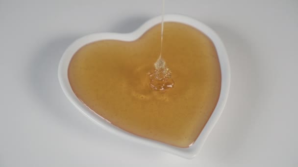A thin stream of bee honey flows in a white plate in the shape of a heart on a white wooden table. Filling. Side view. Natural dessert - Footage, Video