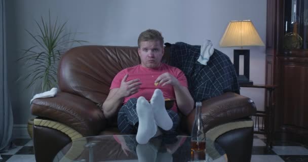 Portrait of lonely Caucasian man chewing chips and watching football on TV. Adult single guy resting alone at home. Loneliness, lifestyle, laziness. Cinema 4k ProRes HQ. - Materiał filmowy, wideo
