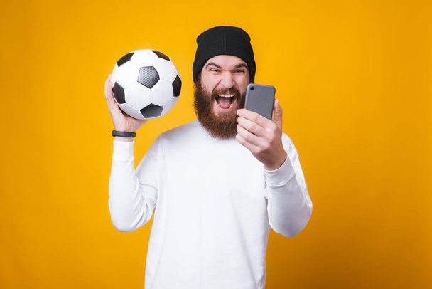 Young bearded man is taking a selfie and holding a soccer ball near yellow wall. - Foto, afbeelding