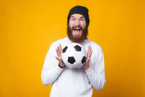 Счастливый бородатый держит взволнованный футбольный мяч на желтой стене
. - Фото, изображение