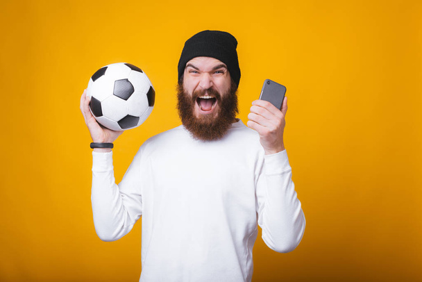 Genç adam bağırıyor, elinde telefon ve sarı duvarın yanında bir futbol topu tutuyor.. - Fotoğraf, Görsel