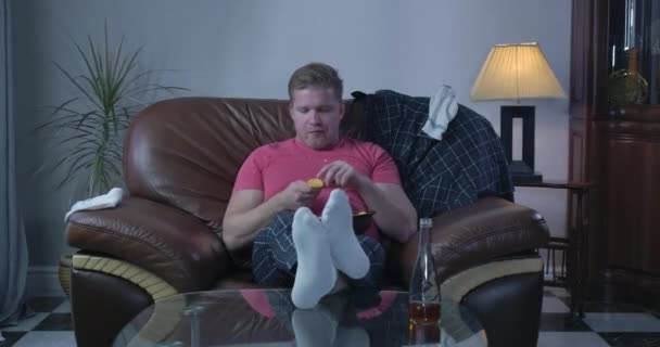 Lonely Caucasian man sitting on sofa and throwing up chips. Young single guy watching TV alone at home. Loneliness, lifestyle, laziness. Cinema 4k ProRes HQ. - Кадры, видео