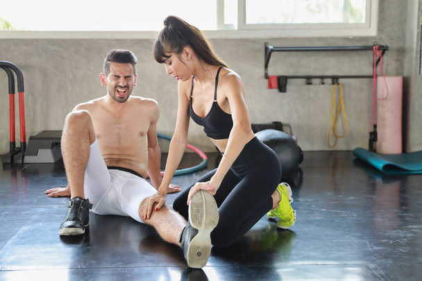 junge kaukasische Frau hilft Fitness-Mann, der Sportanzug trägt und sich durch zu viel Bewegung im Fitnessstudio am Knöchel verletzt - Foto, Bild
