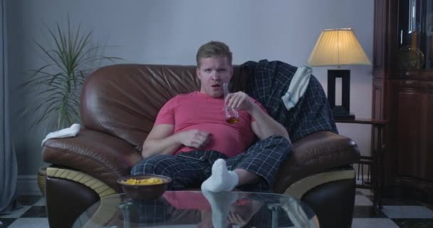 Portrait of lazy adult Caucasian man drinking beer, stretching belly, drinking beer and eating chips. Single guy watching TV alone indoors. Addiction, loneliness, alcoholism. Cinema 4k ProRes HQ. - Materiaali, video