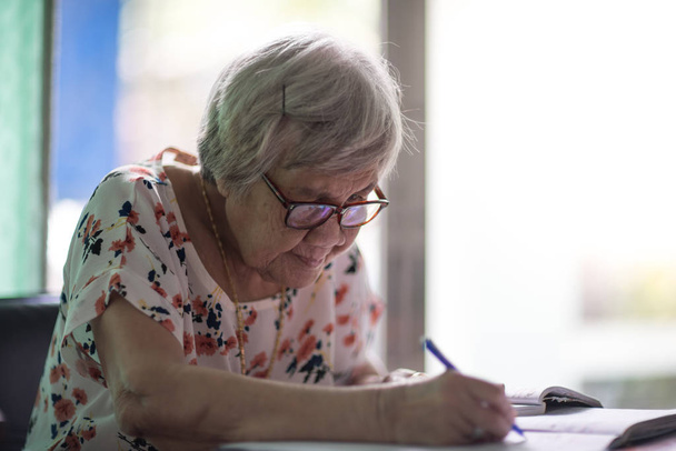 Aasialainen vanha nainen istuu ja kirjoittaa pöydälle lasin wi
 - Valokuva, kuva
