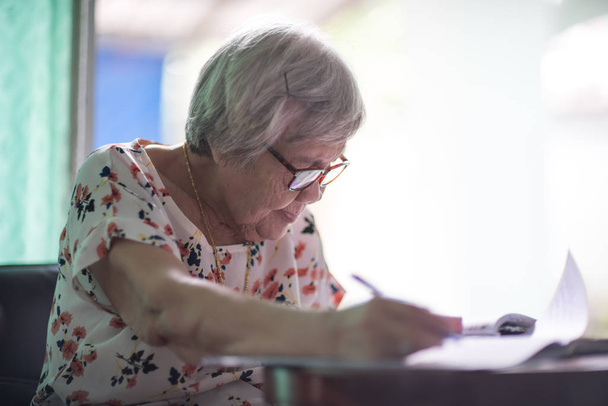 Азиатская старуха сидит и пишет на столе у стеклянной палочки
 - Фото, изображение