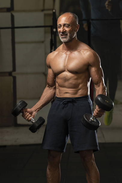 Muscular sem camisa maduro mais velho atleta de musculação com cabelos grisalhos calvos faz um halteres bicep curl em um ginásio mostrando seus abdominais seis-pack
 - Foto, Imagem