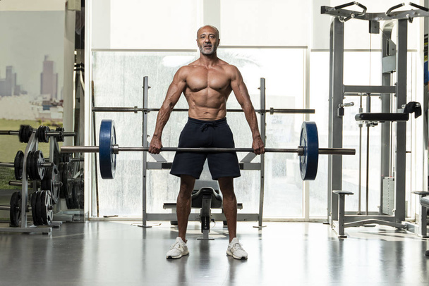 Um forte muscular sem camisa maduro atleta de musculação mais velho com cabelo grisalho careca levantando uma barra pesada, olhando para a câmera em um ginásio
 - Foto, Imagem
