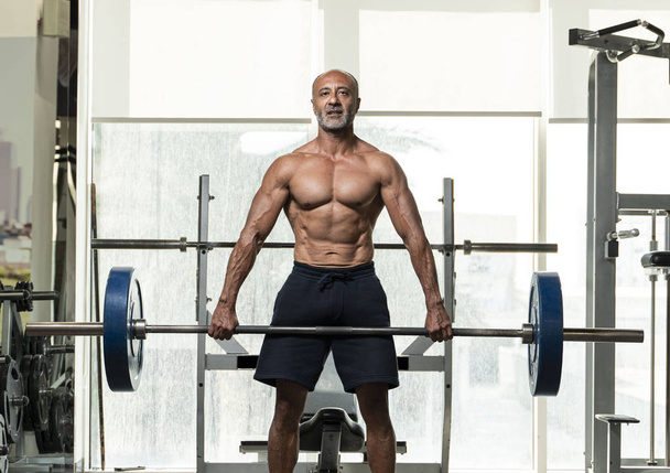 Un atleta mayor maduro musculoso fuerte del levantamiento de pesas sin camisa con el pelo gris calvo que levanta una barra pesada, mirando la cámara en un gimnasio
 - Foto, Imagen