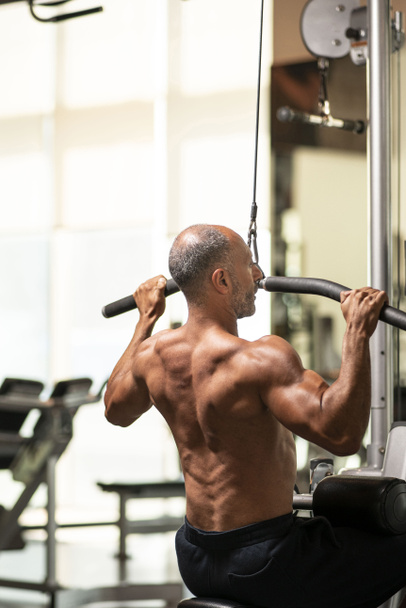 Muscular sem camisa maduro mais velho atleta de musculação com cabelos grisalhos calvos realizando um exercício de remo sentado em uma máquina em um ginásio
 - Foto, Imagem