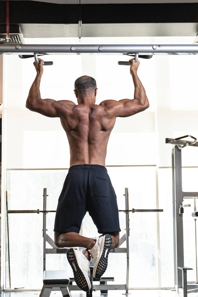 Πλάτη άποψη ενός ισχυρού μυϊκή shirtless ώριμος μεγαλύτερος αθλητής bodybuilding με φαλάκρα γκρίζα μαλλιά εκτελεί ένα pull-up άσκηση σε ένα γυμναστήριο - Φωτογραφία, εικόνα