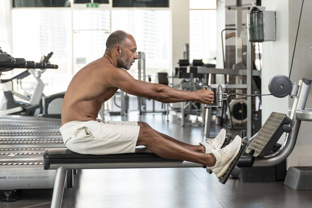 Muscular sem camisa maduro mais velho atleta de musculação com cabelos grisalhos calvos realizando um exercício de remo sentado em uma máquina em um ginásio
 - Foto, Imagem