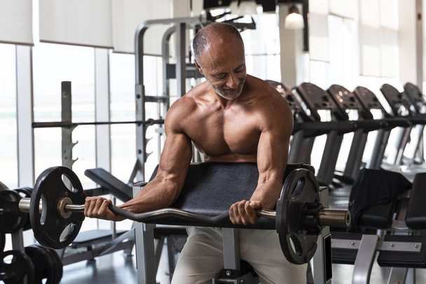 Gespierde shirtloze volwassen oudere bodybuilding atleet met kalende grijze haren doet een lange biceps krullen in een fitnessruimte - Foto, afbeelding