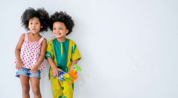 zwei kleine afrikanische Jungen und Mädchen stehen vor einer weißen Wand und drücken unterschiedliche Aktionen aus, und sie sehen entspannt und lustig aus, wenn ein Junge ein paar Spielzeuge in der Hand hält. - Foto, Bild
