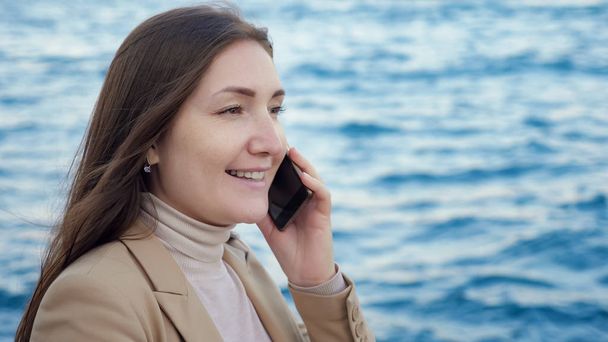 lady talks on phone standing on coast against blurry sea - Φωτογραφία, εικόνα