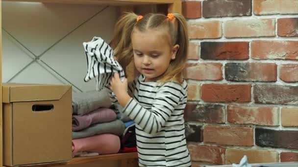 Το κοριτσάκι τυλίγει τα ρούχα σε ένα χαρτόκουτο στο σπίτι. - Πλάνα, βίντεο