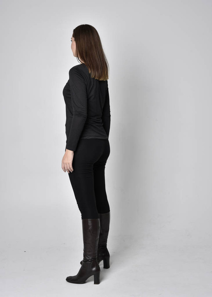 full length portrait of a pretty brunette girl wearing a black shirt leather boots. Standing pose, facing away from the camera,  on a grey studio background. - Foto, afbeelding