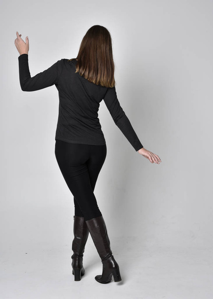 full length portrait of a pretty brunette girl wearing a black shirt leather boots. Standing pose, facing away from the camera,  on a grey studio background. - Foto, afbeelding