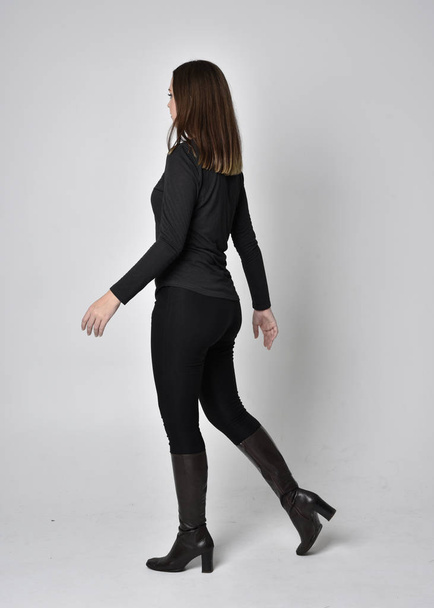 full length portrait of a pretty brunette girl wearing a black shirt leather boots. Standing pose, facing away from the camera,  on a grey studio background. - Photo, image
