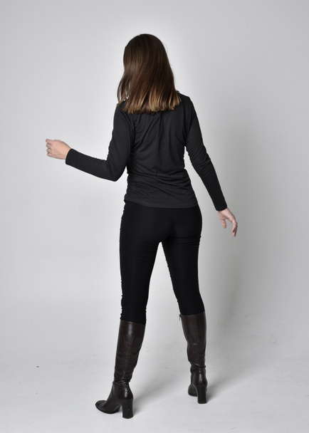 full length portrait of a pretty brunette girl wearing a black shirt leather boots. Standing pose, facing away from the camera,  on a grey studio background. - Foto, immagini