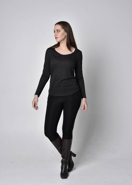 full length portrait of a pretty brunette girl wearing a black shirt leather boots. Standing pose, facing towards the camera,  on a grey studio background. - Photo, Image