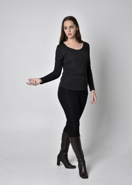 full length portrait of a pretty brunette girl wearing a black shirt leather boots. Standing pose, facing towards the camera,  on a grey studio background. - Foto, imagen