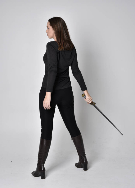 full length portrait of a pretty brunette girl wearing a black shirt and leather boots, holding a sword. Standing pose facing away and holding a sword,  on a grey studio background. - Fotó, kép