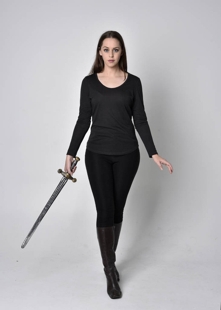 黒いシャツと革のブーツを着た美しいブルネットの女の子の完全な長さの肖像画、剣を持っています。立ちポーズ剣を持ってグレーのスタジオの背景に. - 写真・画像