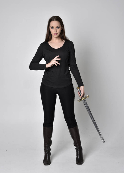 πορτραίτο ενός όμορφου μελαχρινού κοριτσιού με μαύρο πουκάμισο και δερμάτινες μπότες, κρατώντας ένα σπαθί. Μόνιμη στάση, κρατώντας ένα σπαθί, σε ένα γκρι φόντο στούντιο. - Φωτογραφία, εικόνα