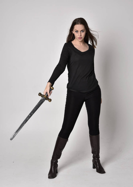 Ganzkörperporträt eines hübschen, brünetten Mädchens in schwarzem Hemd und Lederstiefeln, das ein Schwert in der Hand hält. Stehende Pose, ein Schwert in der Hand, auf grauem Studiohintergrund. - Foto, Bild