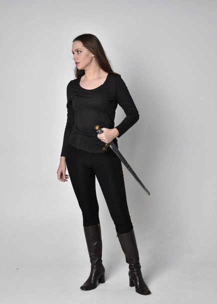 full length portrait of a pretty brunette girl wearing a black shirt and leather boots, holding a sword. Standing pose, holding a sword, on a grey studio background. - Fotografie, Obrázek