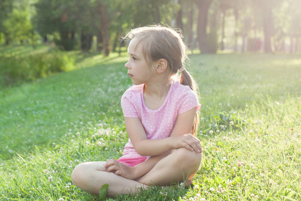 Beautiful little girl in pink t-shirt on grass. Tired girl after walk in the city park - 写真・画像