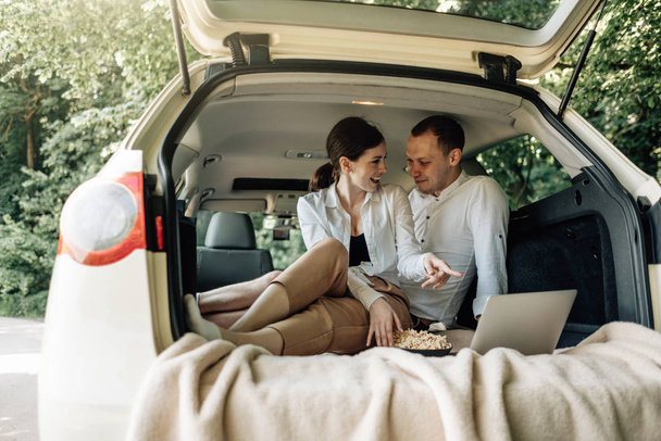 Jovem casal feliz vestido de igual em T-shirt branca Sentado no tronco do carro com laptop e pipoca na beira da estrada, fim de semana fora da cidade, feriados e Road Trip Concept
 - Foto, Imagem