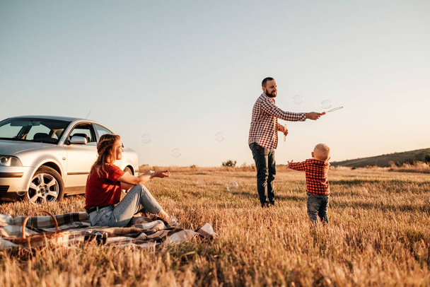 Heureux jeune famille maman et papa avec leur petit fils profiter week-end d'été pique-nique sur la voiture en dehors de la ville, jouer avec des bulles
 - Photo, image