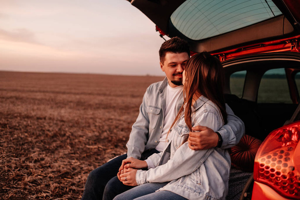 Jovem casal feliz vestido igual em camisa branca e jeans sentado em seu novo tronco de carro, belo pôr do sol no campo, férias e conceito de viagem
 - Foto, Imagem