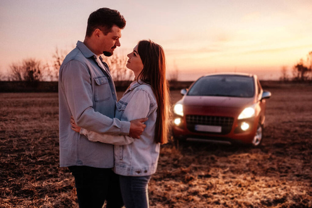 Young Happy Couple Dressed Alike in White Shirt and Jeans Enjoying Road Trip at Their New Car, Beautiful Sunset on the Field, Vacation and Travel Concept - Foto, afbeelding