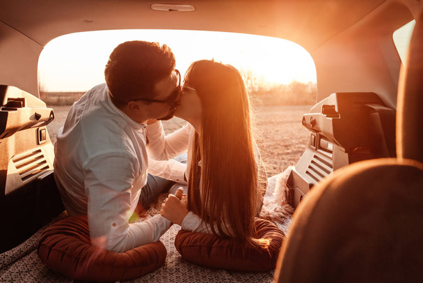 白いシャツを着た若い幸せなカップルと新しい車でロードトリップを楽しむジーンズ、フィールドでの美しい夕日、休暇と旅行のコンセプト - 写真・画像