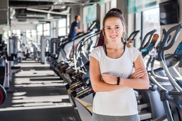 Smiling sports caucasian woman standing with arms cross and looking camera at fitness gym background with copy space in left. Trainer or instructor fitness coach concept - Photo, Image