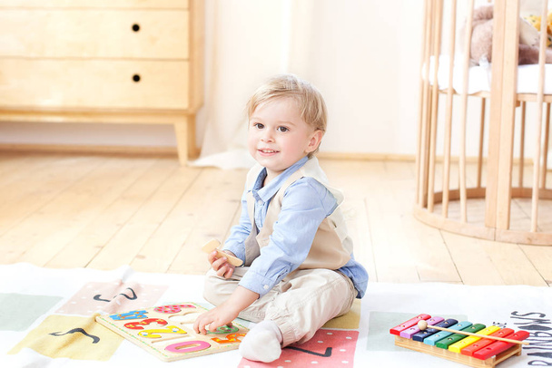 Chlapče. Chlapec si hraje s dřevěnými hračkami a čísly. Vzdělávací dřevěné hračky pro dítě. Portrét chlapce sedícího na podlaze v dětském pokoji ve skandinávském stylu. Eko hračky, dekor  - Fotografie, Obrázek