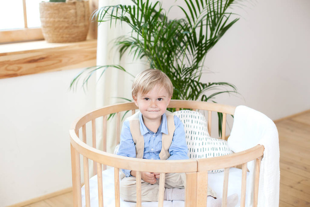 little boy. Portrait of a happy boy playing in a baby cot. The boy sits alone in a crib in the nursery. Lonely baby stay in the crib. Waif child. The child in bed is smiling. Interior children's room. - Photo, Image