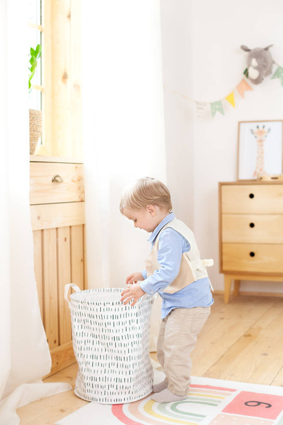 A little boy puts toys in a Scandinavian basket for a children's room. Eco-friendly decor child's room. Portrait of a boy playing in kindergarten. Kids room and interior design. Baby boy at home. - Fotografie, Obrázek