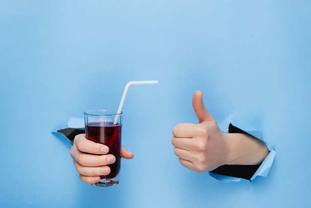 Female hand holding a glass of fresh red juice with a straw through torn blue paper wall. Fruit drinks and healthy nutrition. Hand sign. Hole in wall. Like gesture - Foto, Imagen