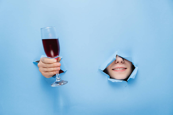 close-up of female nose and lips and a broad smile keeps a glass of red wine through torn hole in blue paper, copy space for your promotion. Valentine's day and Wine bar, winery, wine tasting concept - Foto, immagini