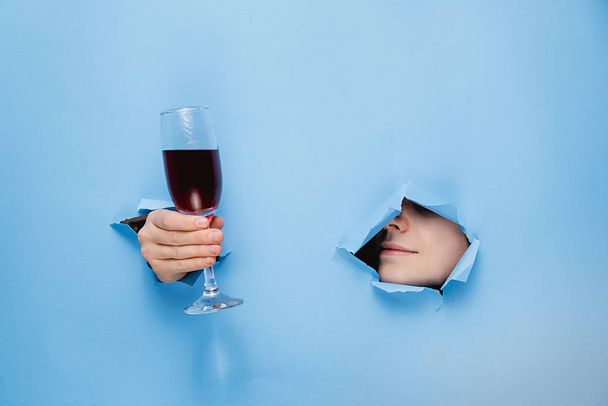 A female nose and lips and hand keeps a glass of red wine through torn hole in blue paper, copy space for your promotion. Valentine's day and Wine bar, winery, wine tasting concept - Fotoğraf, Görsel