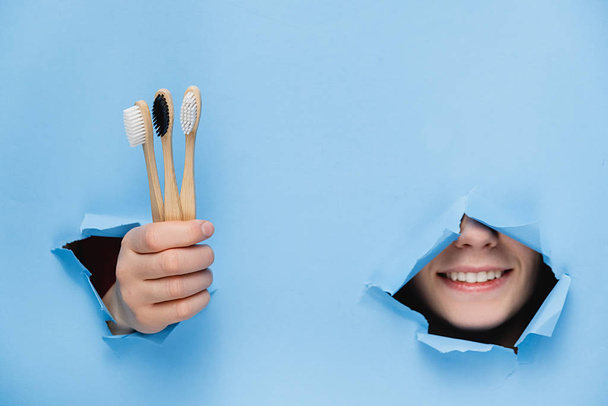 Positive woman with toothy pleasant smile on face, keeps head and bamboo eco toothbrushes through torn hole in blue paper, copy space for your promotion. Dental care and Eco friendly and reuse concept - Foto, afbeelding