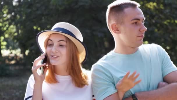 Porträt eines hübschen, positiven Teenagers mit roten Haaren und Strohhut, der fröhlich auf dem Handy redet, während sein Freund gelangweilt neben ihm wartet. - Filmmaterial, Video