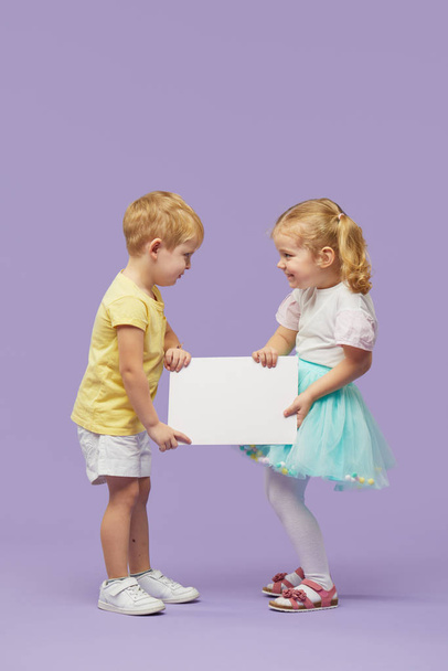 Two little children holding a white banner on purple background. Funny Faces. Copy space for text - 写真・画像