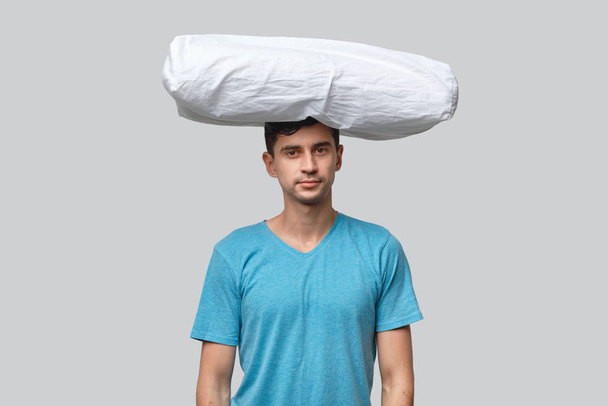 Белая подушка лежит на голове уставшего брюнетки мужчина в синей майке изолированы на сером фоне
. - Фото, изображение