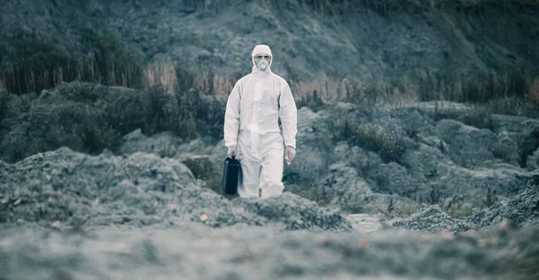 Tecnico di laboratorio in maschera e tuta protettiva chimica, cammina su terreno asciutto con una cassetta degli attrezzi attraverso fumo tossico - Foto, immagini