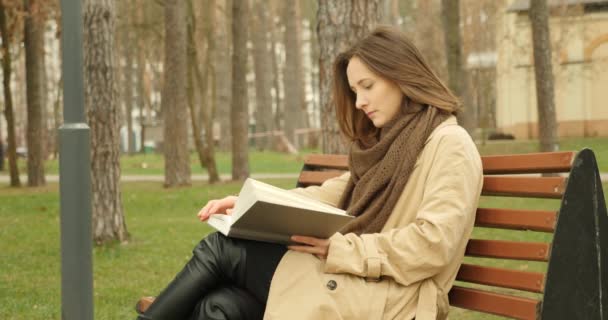 Woman reading book sitting on the bench in autumn park wearing brown trench and scarf. Reading concept - 映像、動画